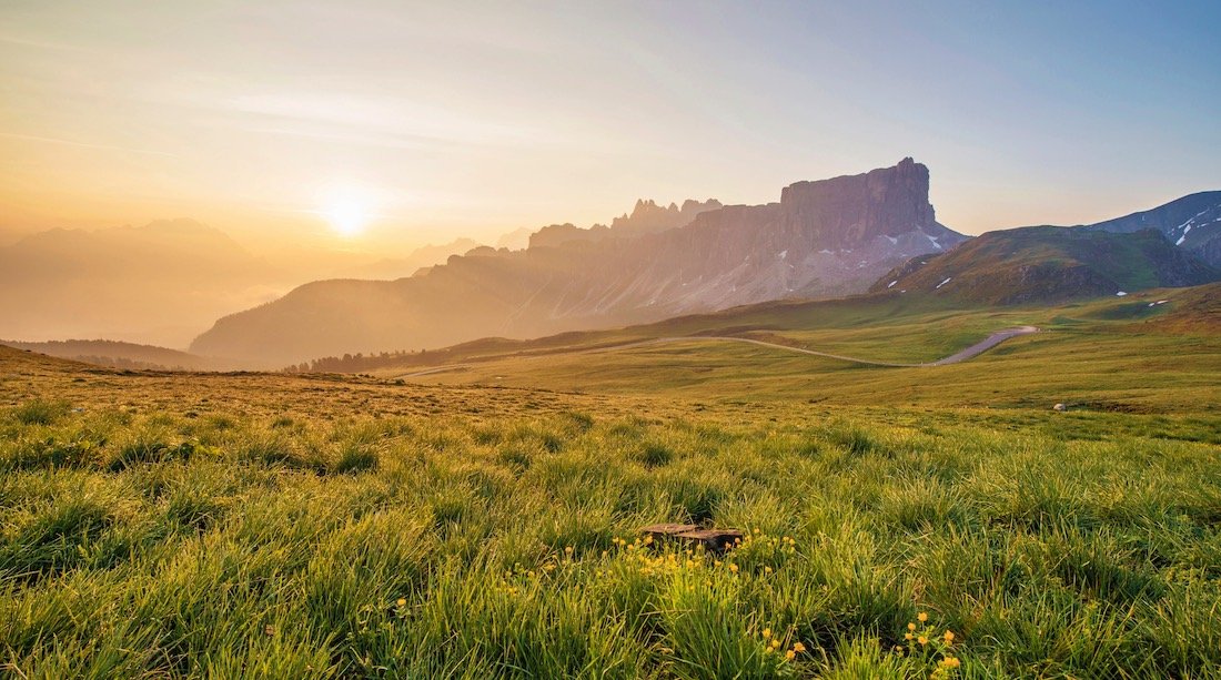 Sun rises over a grassy meadow and rugged mountain peaks