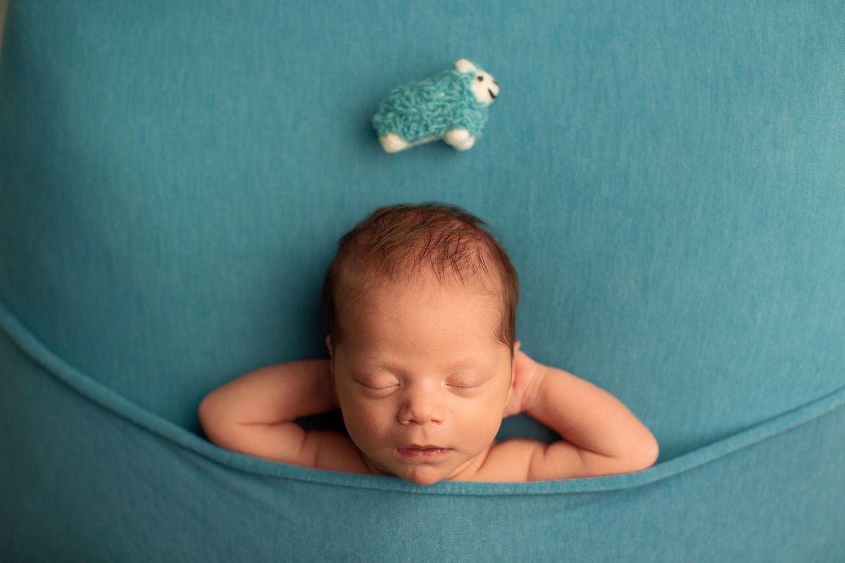Newborn sleeps with hands behind head beneath a toy sheep