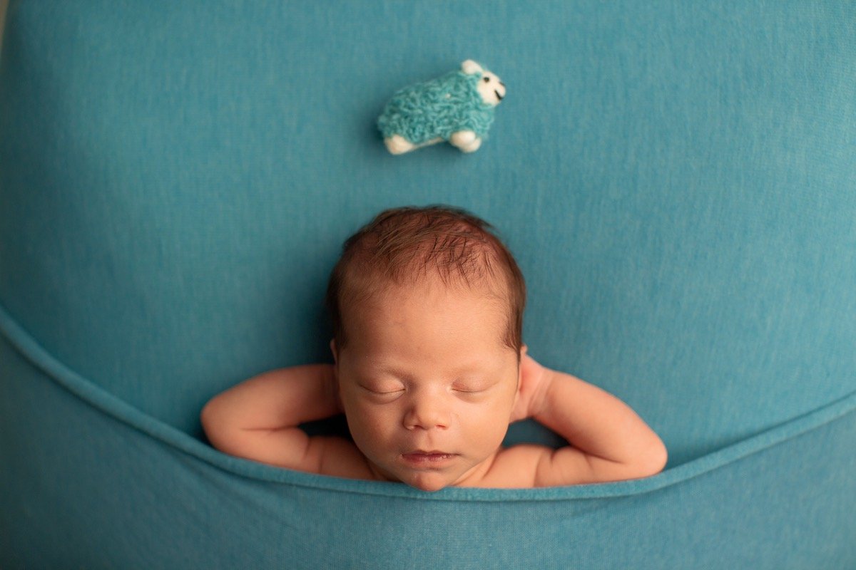 Newborn sleeps with hands behind head beneath a toy sheep