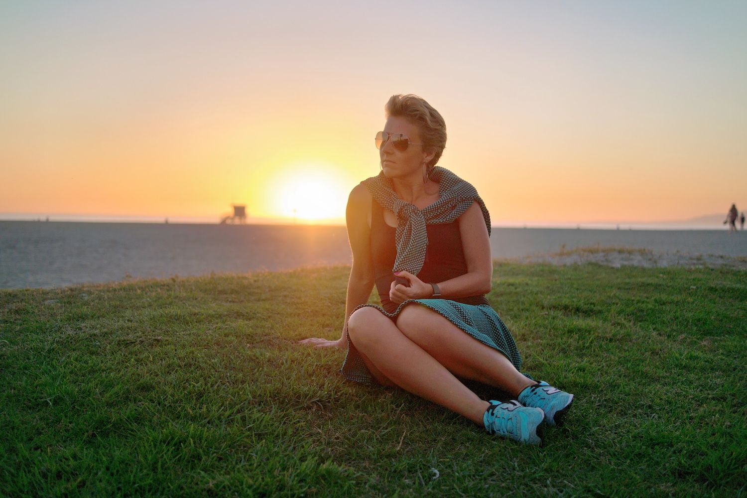 Woman wearing sunglasses sits on grass near a beach at sunset