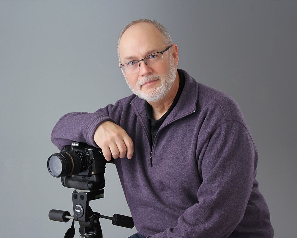 Man with glasses rests his arm on a tripod-mounted camera