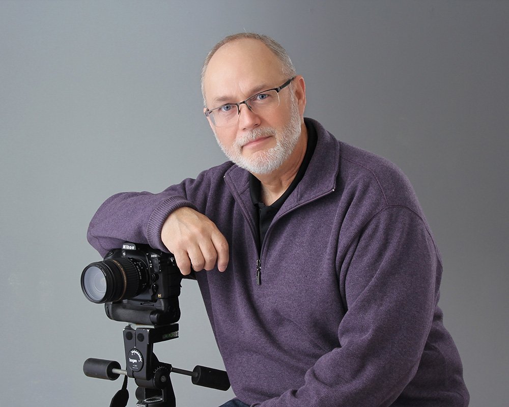 Man with glasses rests his arm on a tripod-mounted camera