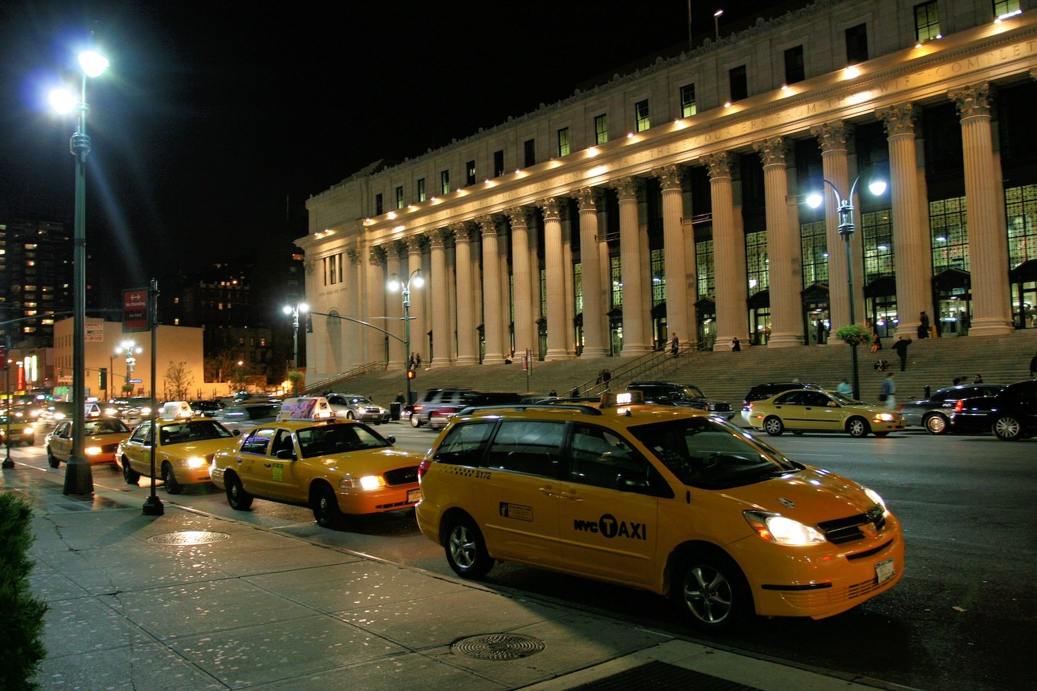 Yellow taxis line a street outside a columned building at night