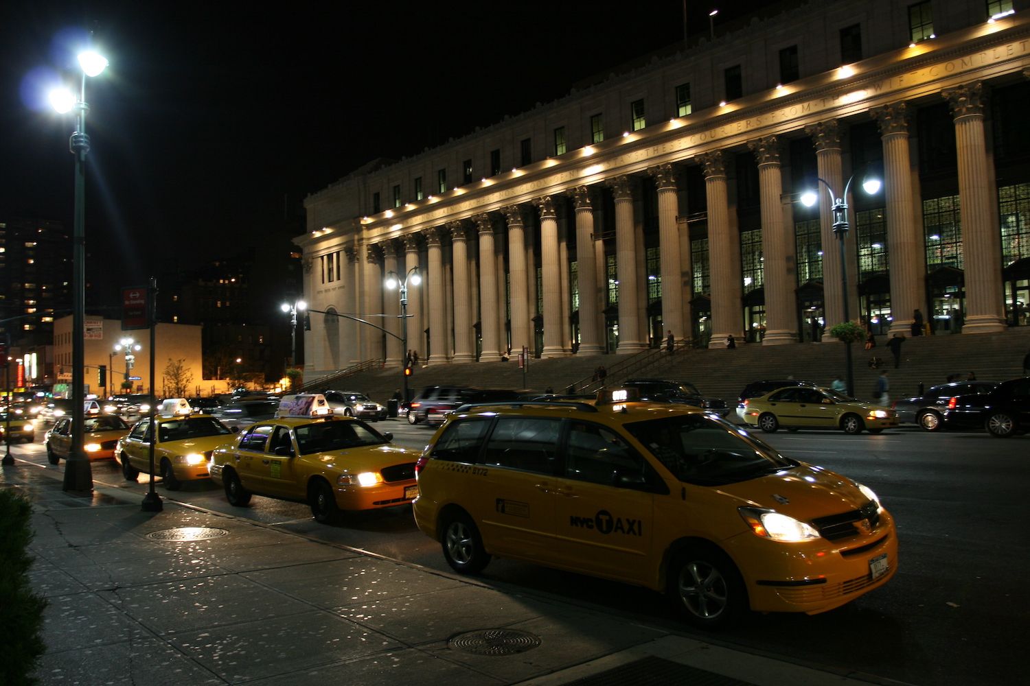 Yellow taxis line a busy street outside a columned building at night