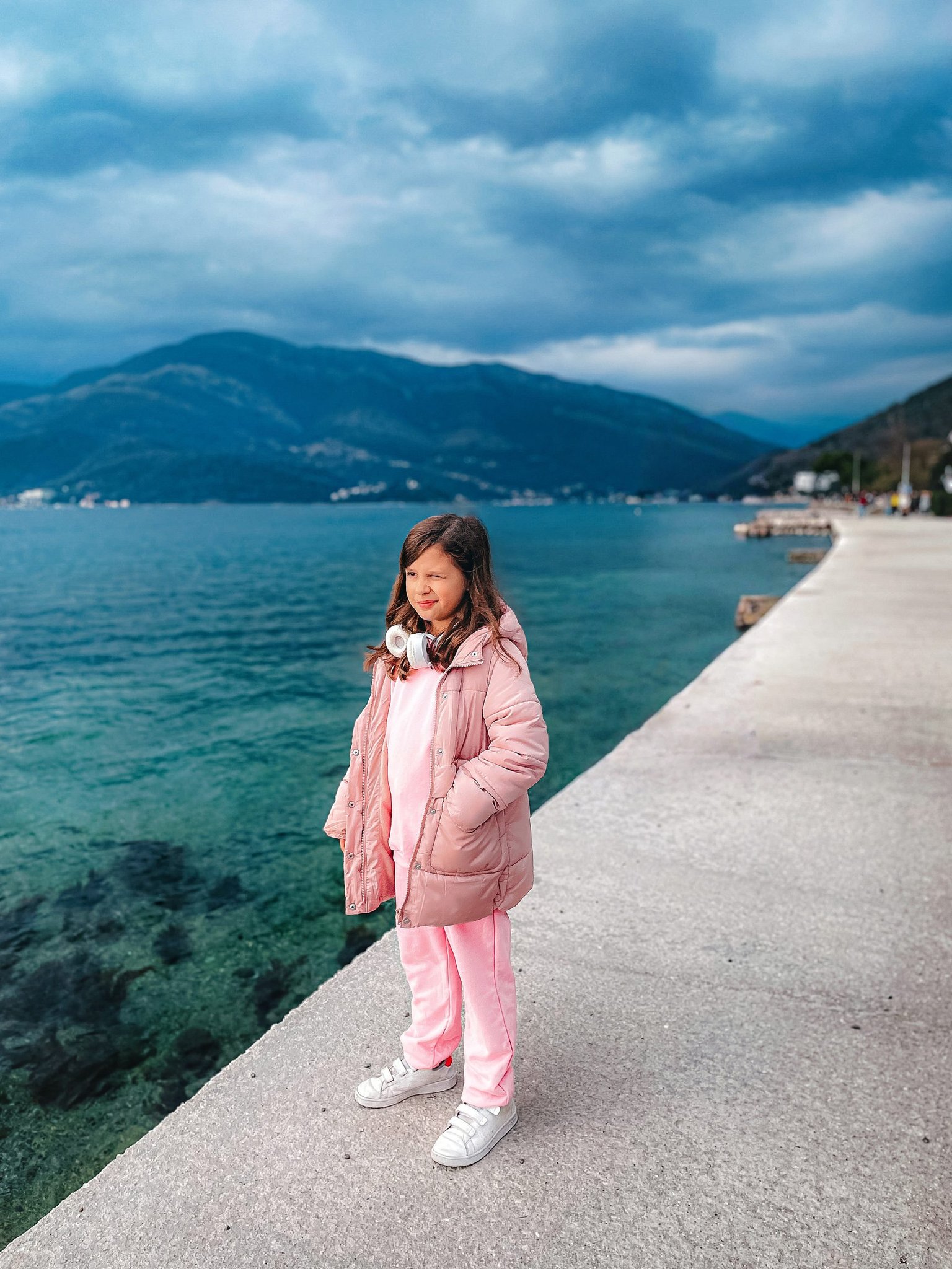 Girl wearing headphones stands on a waterfront pier overlooking mountains