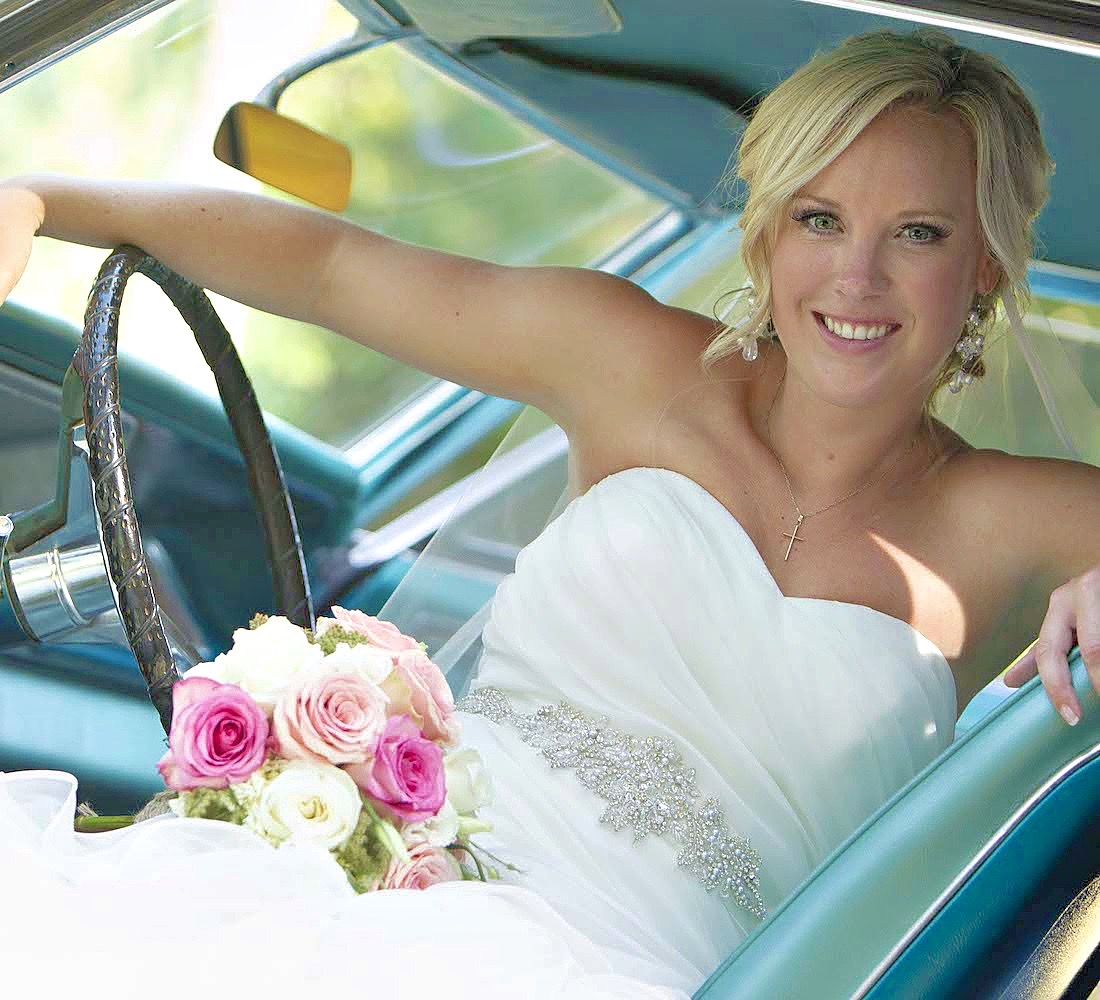 Smiling bride holding a bouquet in the driver's seat of a vintage car