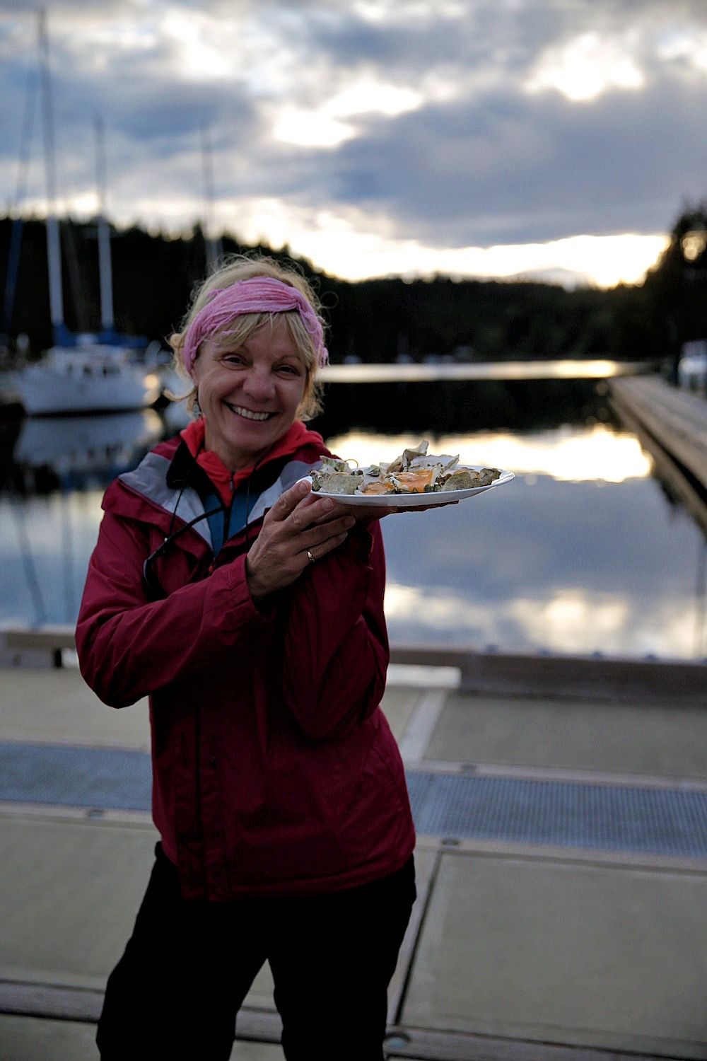 Smiling woman on a marina dock holding a plate of oysters at dusk