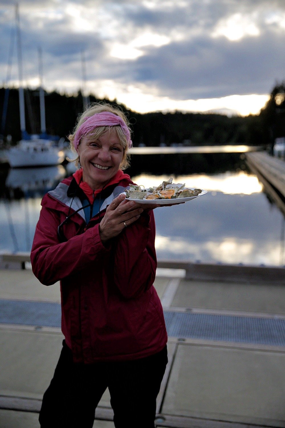 Smiling woman on a marina dock holding a plate of oysters at dusk