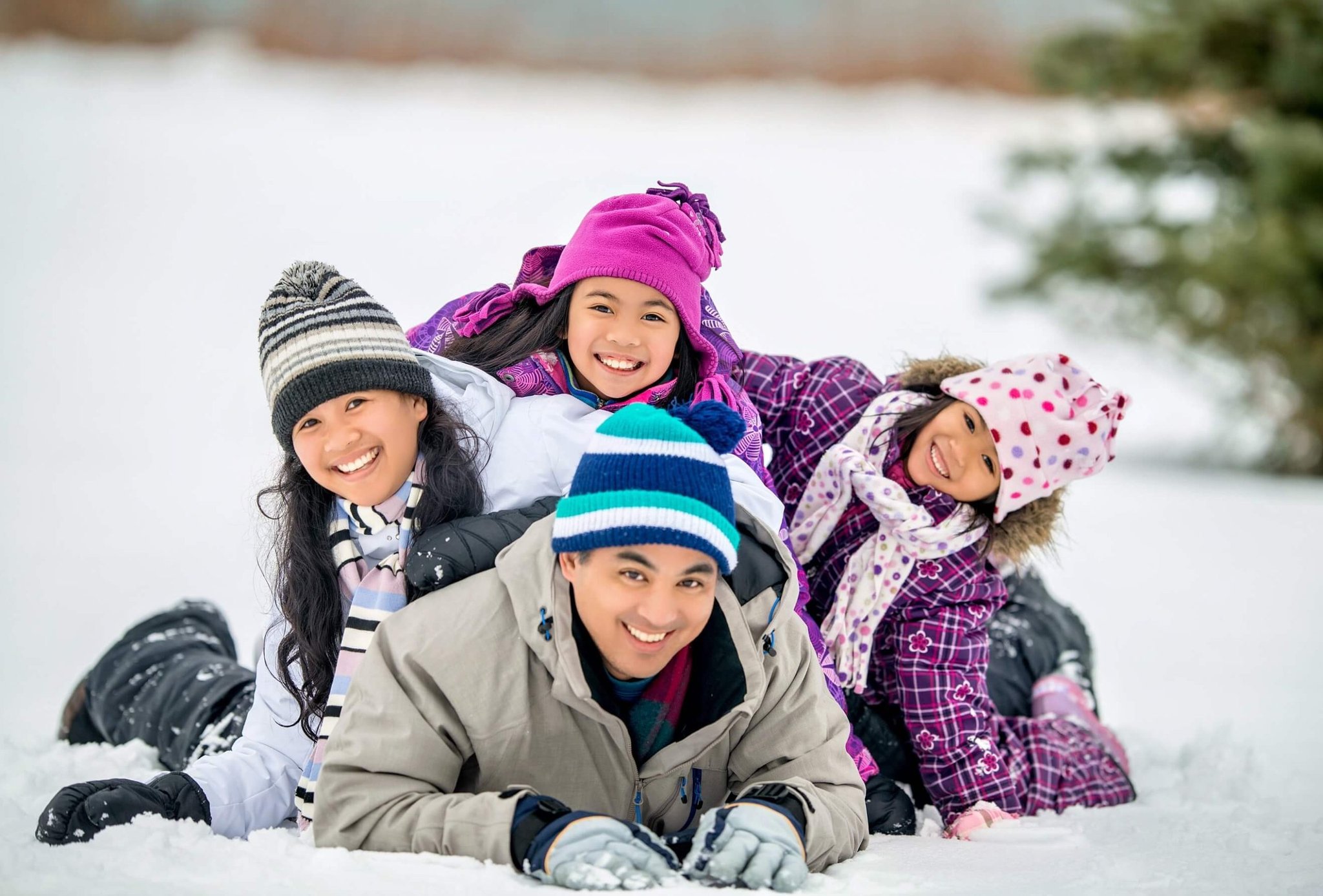 Family of four lying stacked in the snow wearing winter hats and coats