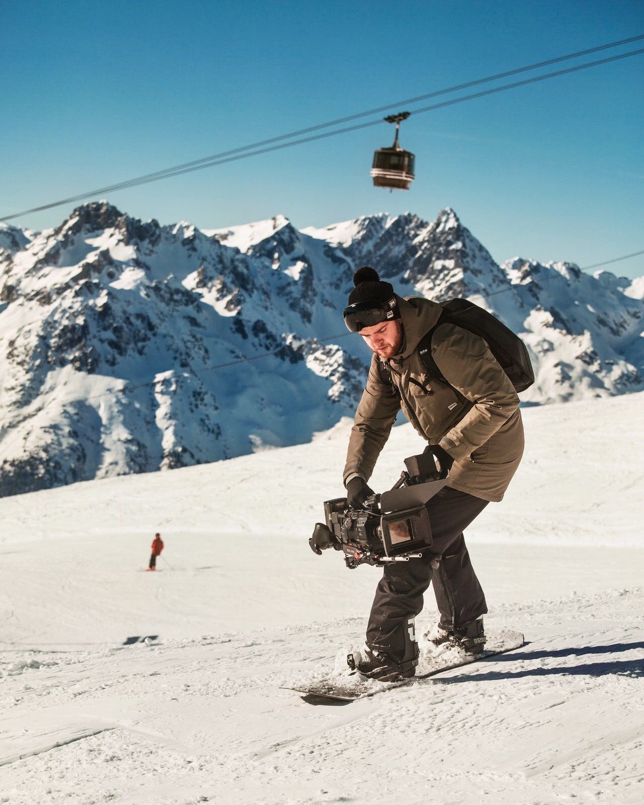 Videographer on a snowboard filming with a cinema camera below a mountain cable car