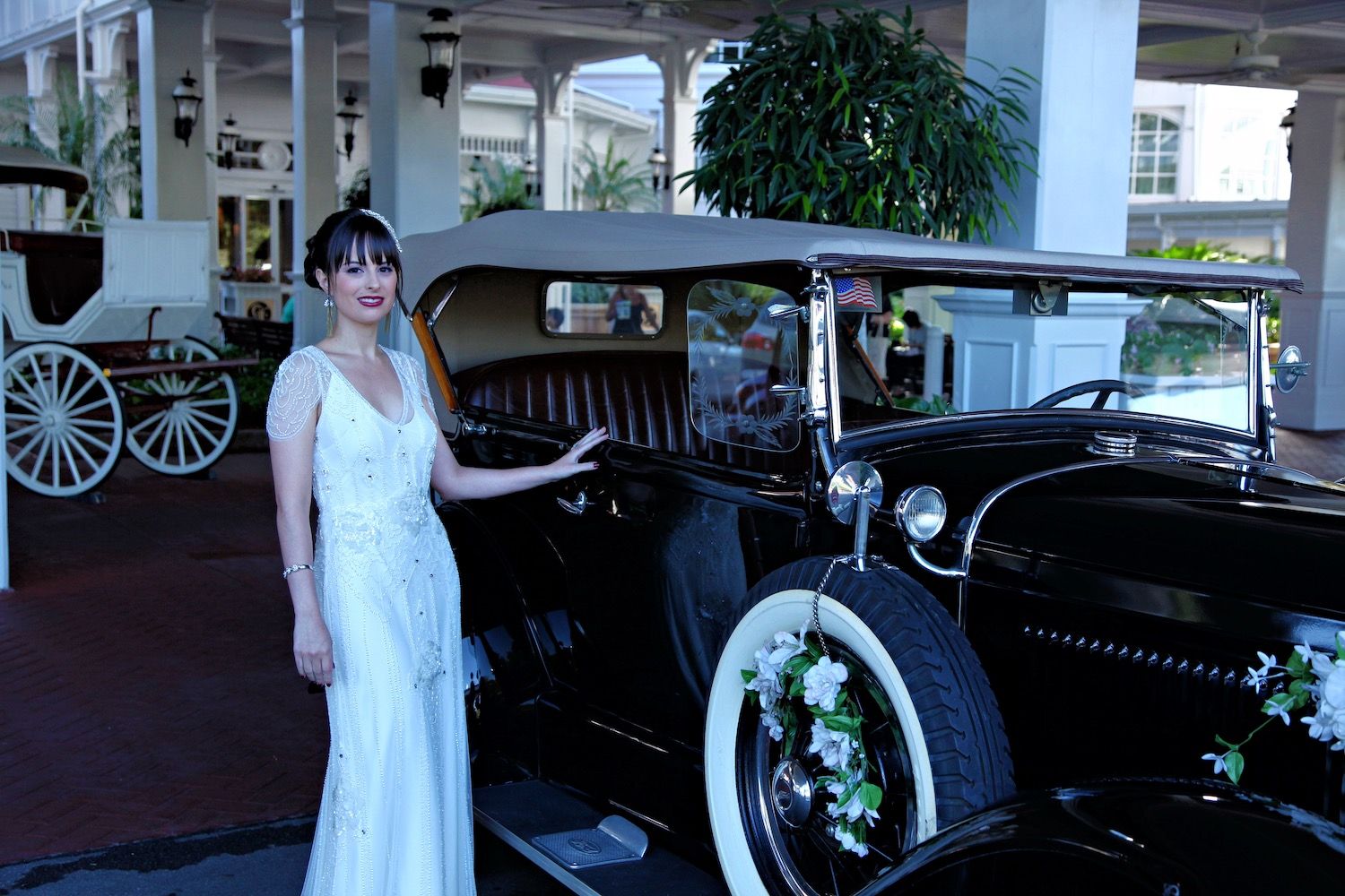 Bride in a white gown standing beside a vintage black car decorated with flowers