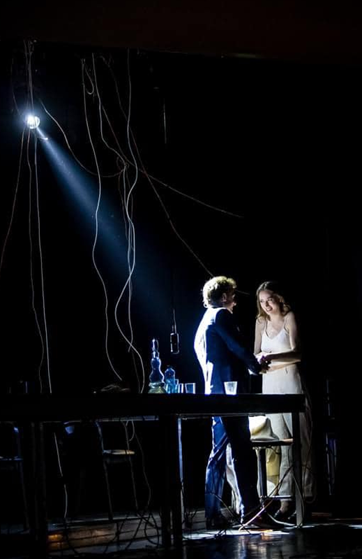 Photo from the performance: a girl is sitting at a table in the spotlight, with twisted cables hanging from the ceiling.