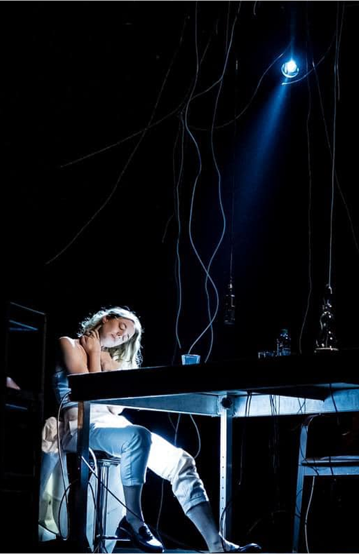 Photo from the performance: a girl sits at a table in the spotlight, twisted cables hanging from the ceiling.