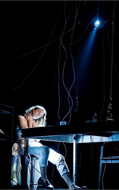Photo from the performance: a girl sits at a table in the spotlight, twisted cables hanging from the ceiling.