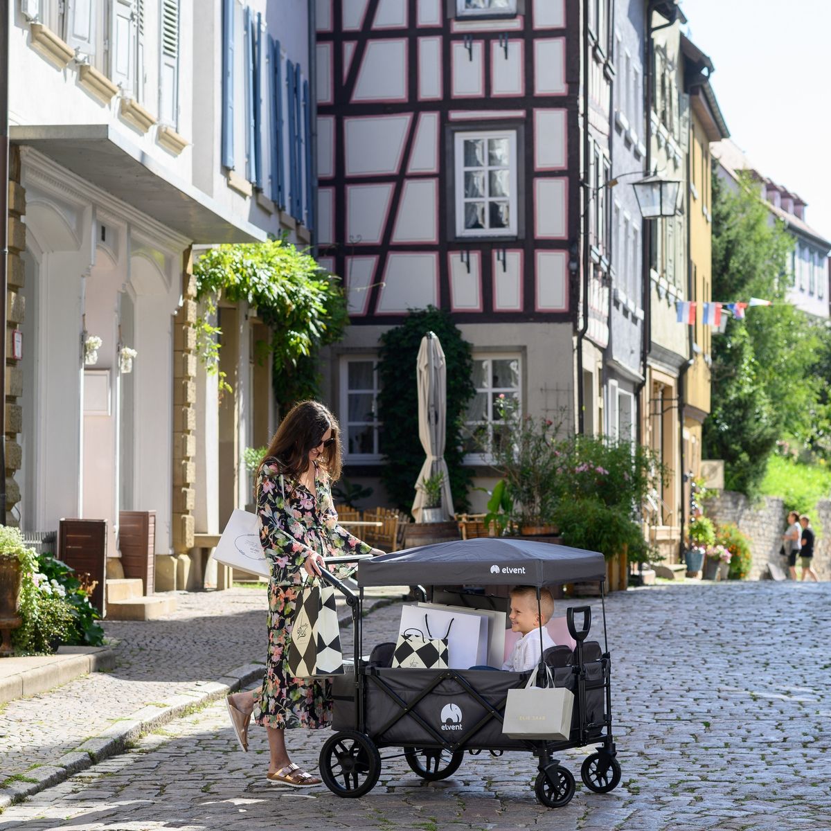 WagonPro City in Indigoblau mit Dach in einer Altstadtgasse, eine Frau mit Einkaufstüten zieht ein Kind