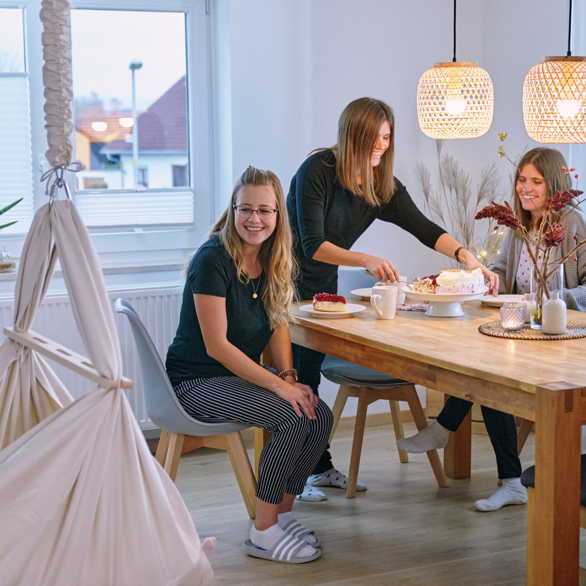 Naturbeige Lullaby Federwiege hängt im Esszimmer neben einem Tisch, an dem drei Frauen Kuchen essen (Mit KI bearbeitetes Bild)