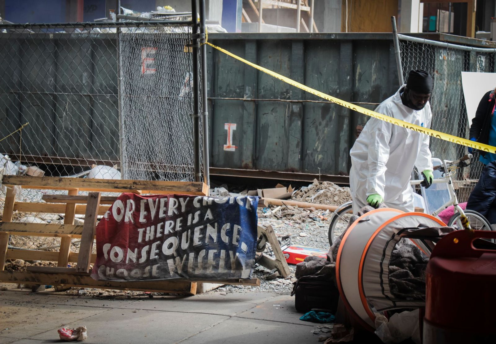 A DC city worker moves belongings past the caution tape as a weathered sign next to him reads “For every choice there is a consequence. Choose wisely.” Eleanor Goldfield | ArtKillingApathy.com