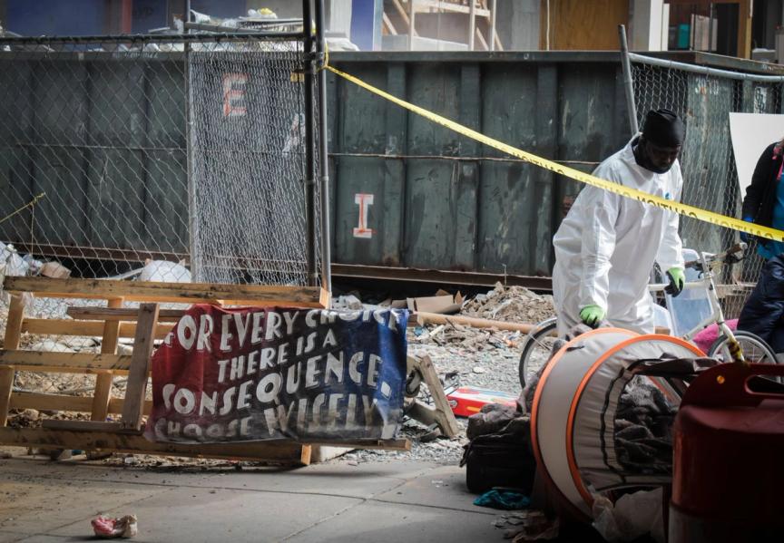 A DC city worker moves belongings past the caution tape as a weathered sign next to him reads “For every choice there is a consequence. Choose wisely.” Eleanor Goldfield | ArtKillingApathy.com