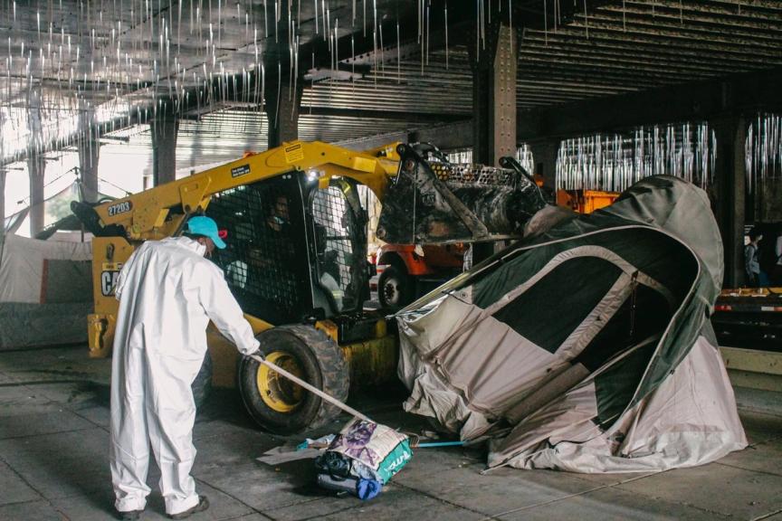 A DC city worker operating a front-end loader crushes a person’s tent and belongings as another city worker pushes sweeps belongings to the side at the M & 2nd St. NE underpass. 