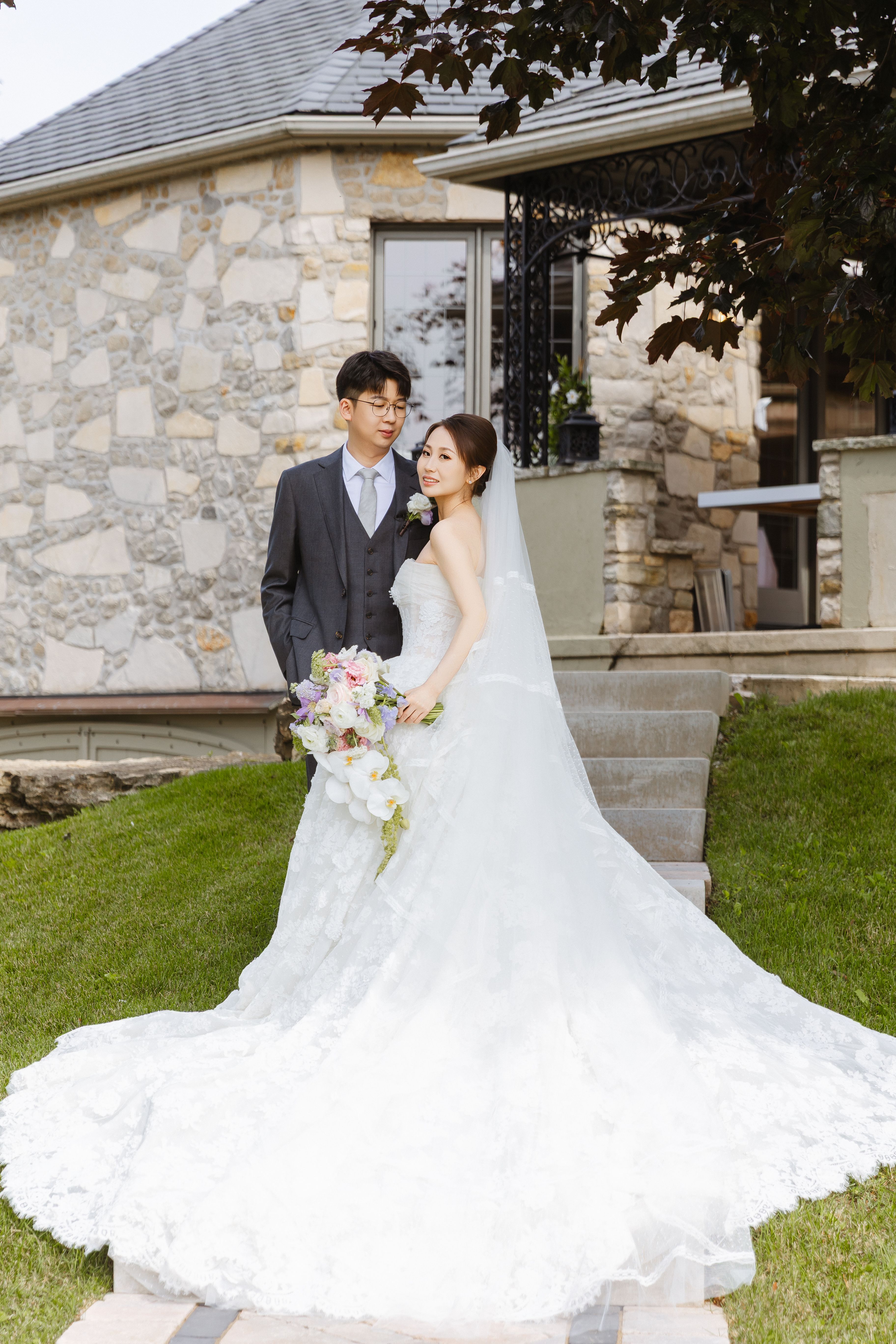 A bride and groom outside the Lincoln Estates manor, with the bride's lace train spread across the steps and lawn.