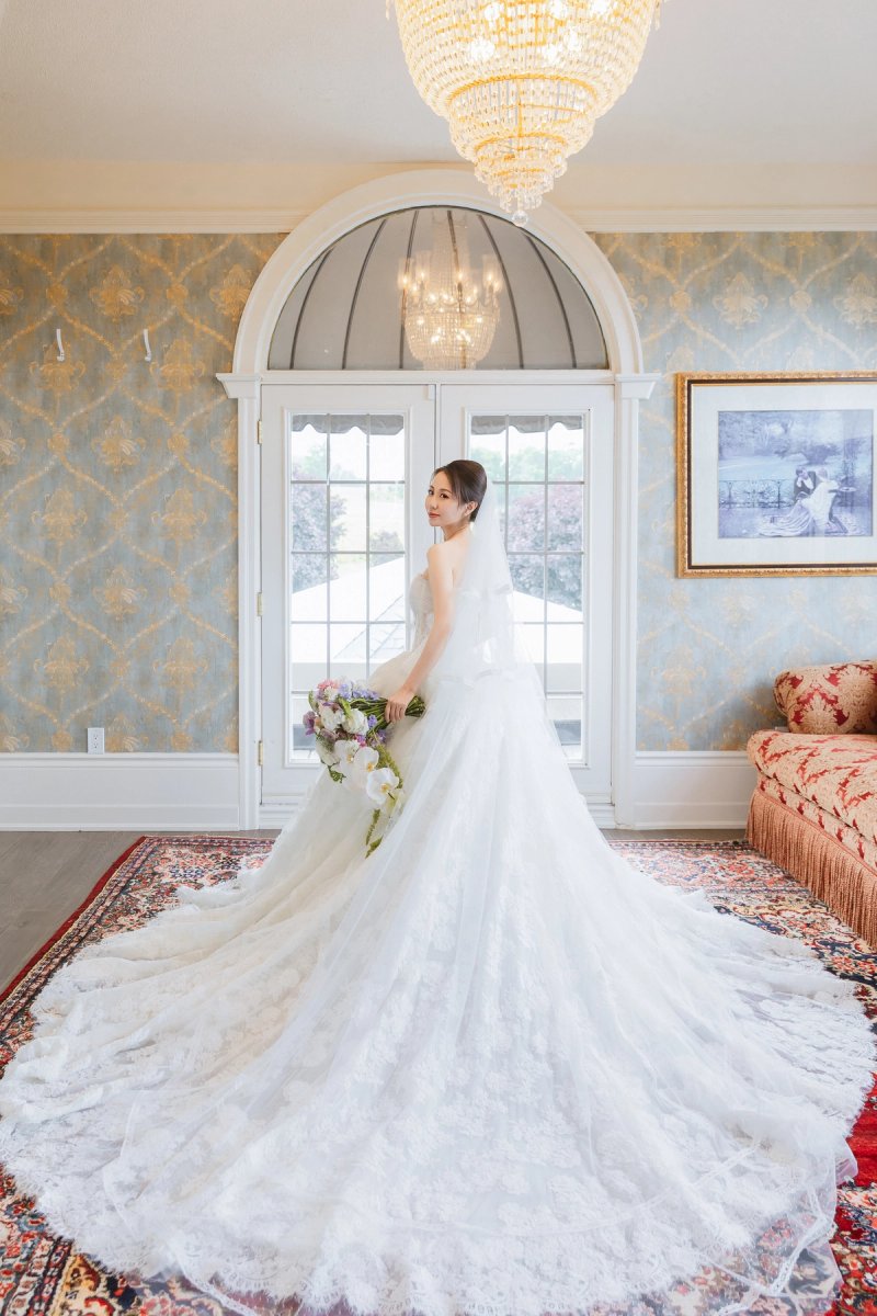 A bride with a long lace train beneath a chandelier, beside arched glass doors and a red upholstered sofa at Lincoln Estates.