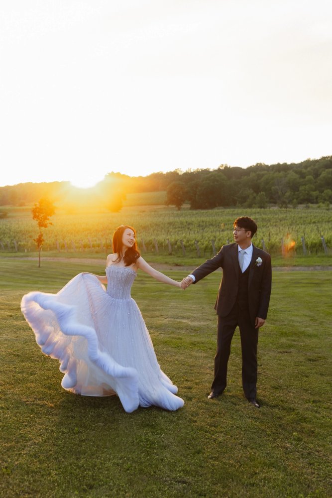A couple holding hands on the Lincoln Estates lawn, the bride lifting a pale blue skirt with the sun low above the vineyard.