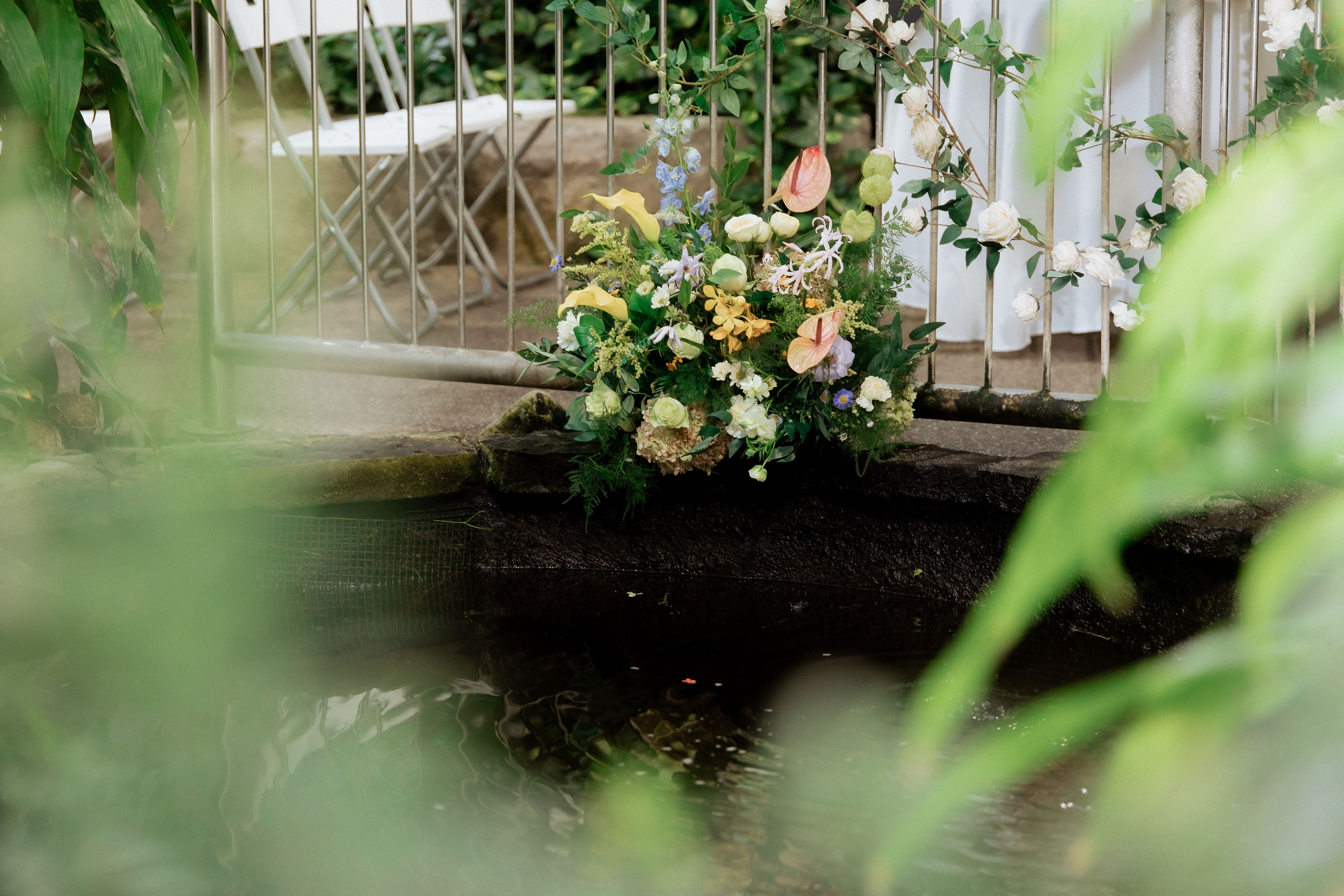 Pastel floral arrangement beside a bridge railing above water, framed by leaves at Cambridge Butterfly Conservatory.
