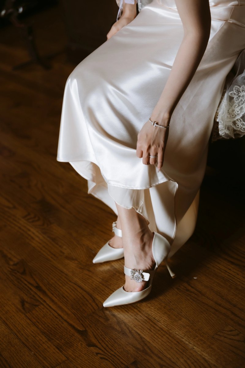 Close-up of the bride's satin dress, ring and white pointed shoes with sparkling buckles on a wooden floor at Casa Loma.