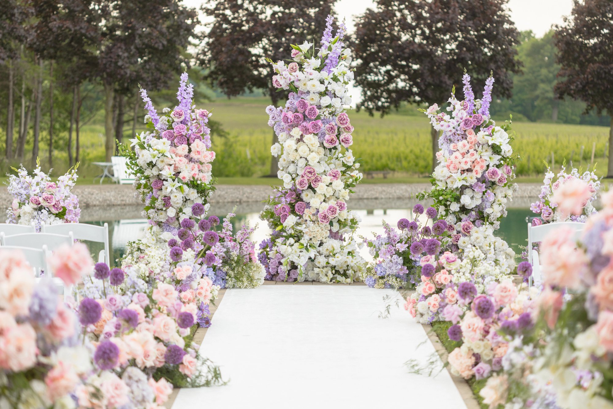 Tall white, pink and purple floral arrangements at the end of a white ceremony aisle beside the pond at Lincoln Estates.