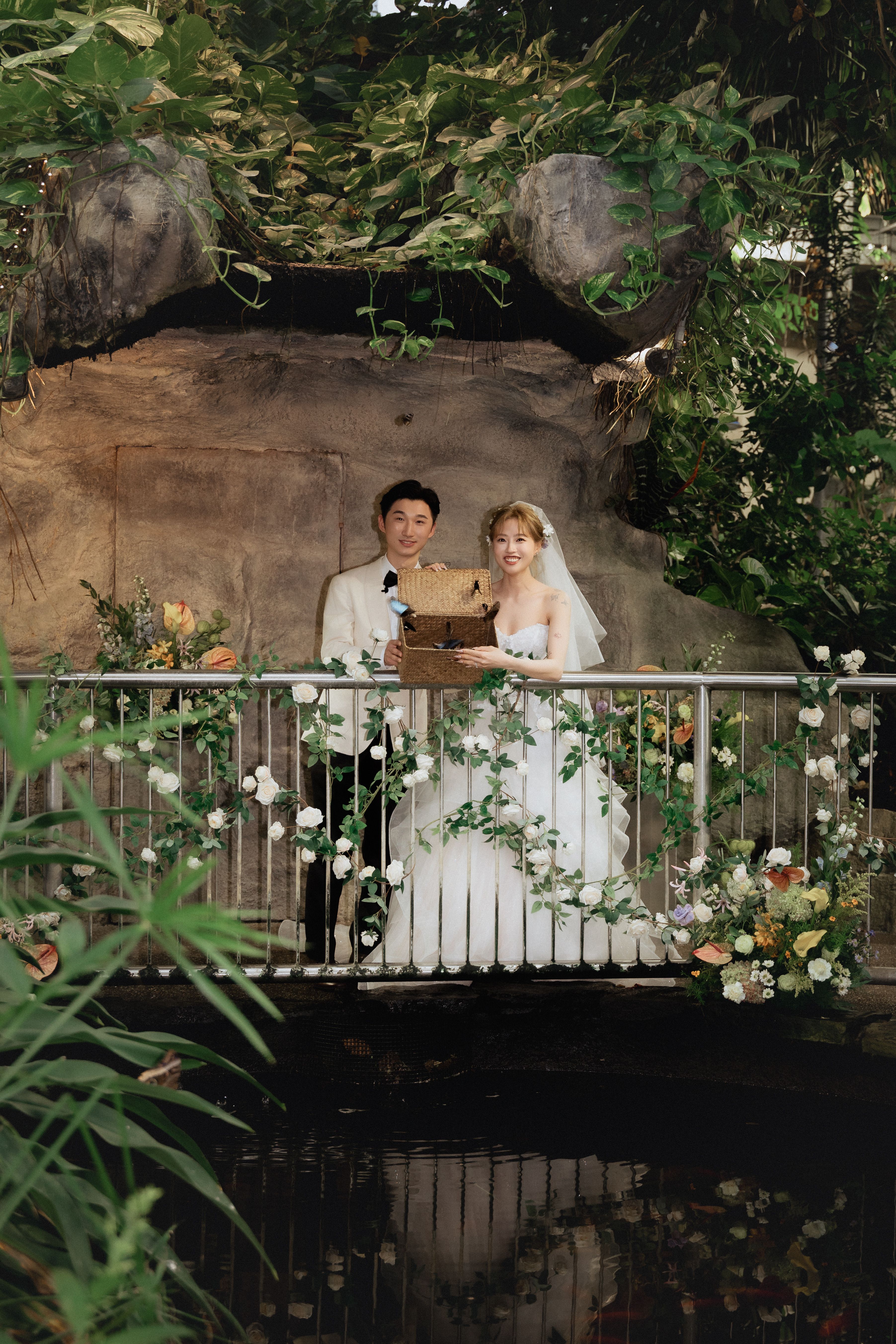Bride and groom holding an open wicker basket on a flower-decorated bridge with butterflies visible at Cambridge Butterfly Conservatory.