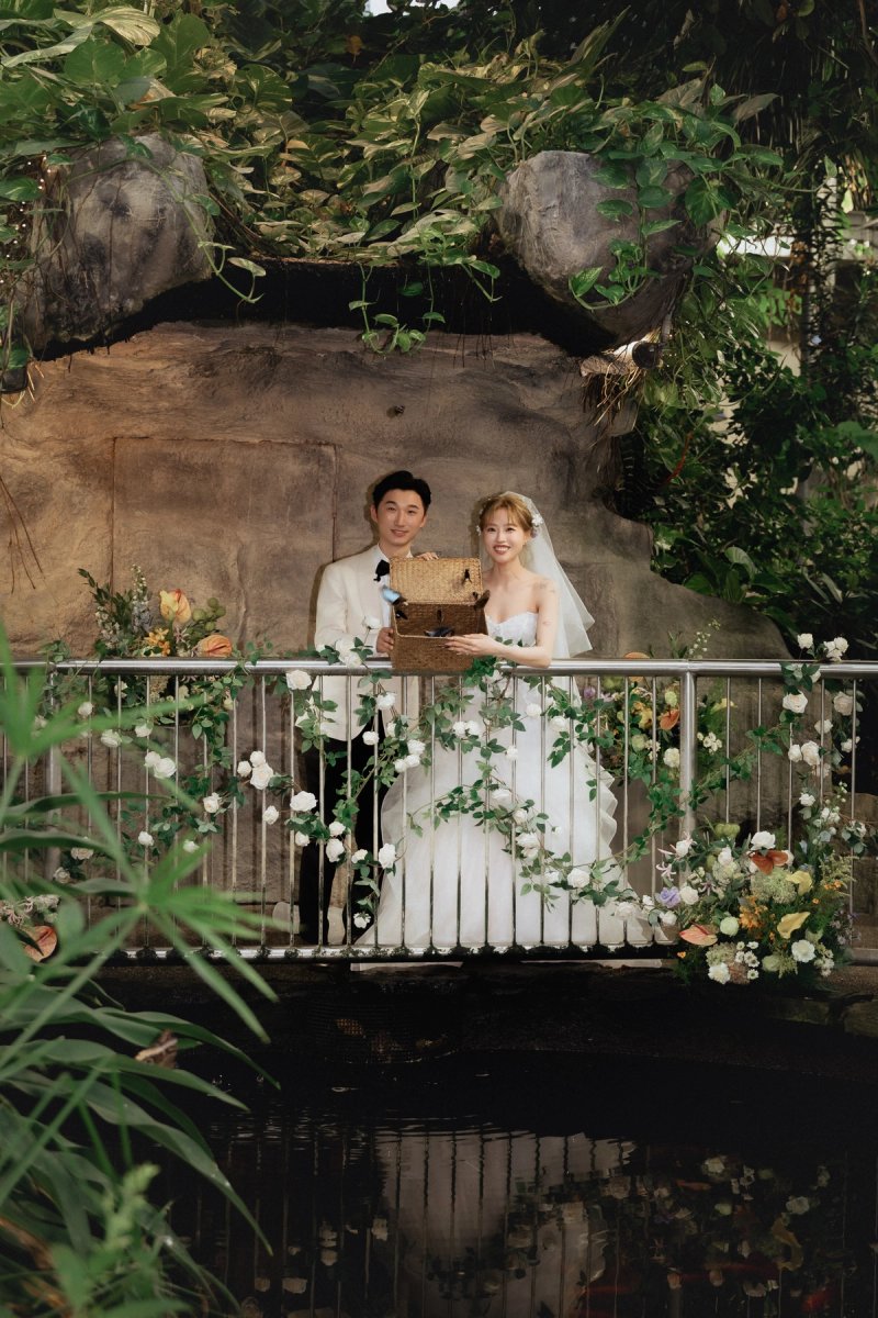 Bride and groom holding an open wicker basket on a flower-decorated bridge with butterflies visible at Cambridge Butterfly Conservatory.