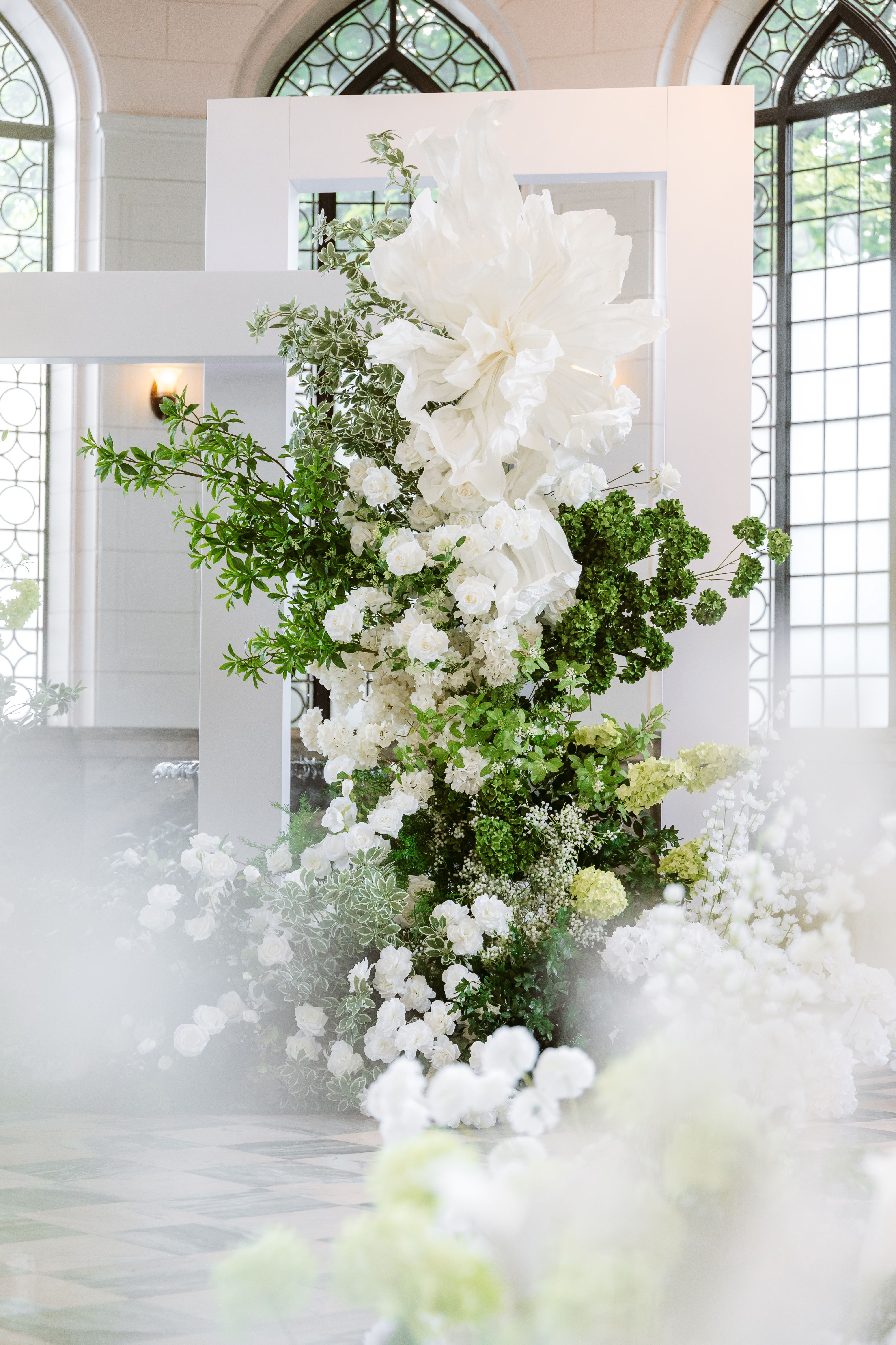 White flowers and leafy greenery surround a white rectangular ceremony frame in front of Casa Loma's arched windows.