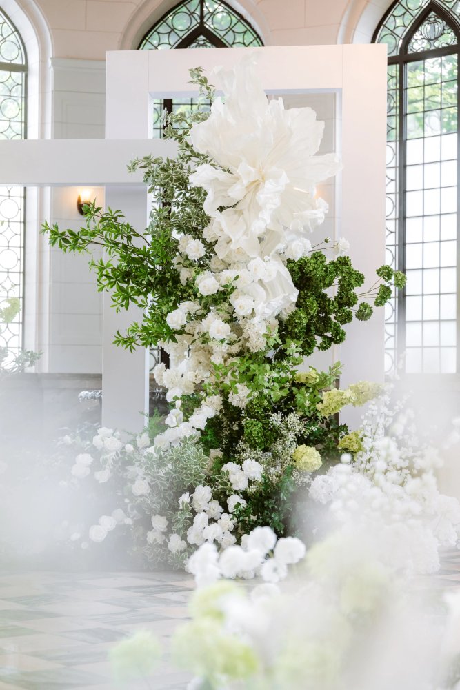 White flowers and leafy greenery surround a white rectangular ceremony frame in front of Casa Loma's arched windows.