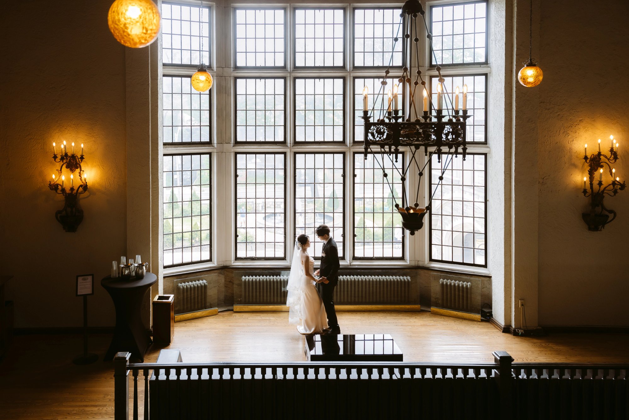 Couple holding hands beneath tall gridded windows and a chandelier at Casa Loma, with a black piano and railing in the foreground.