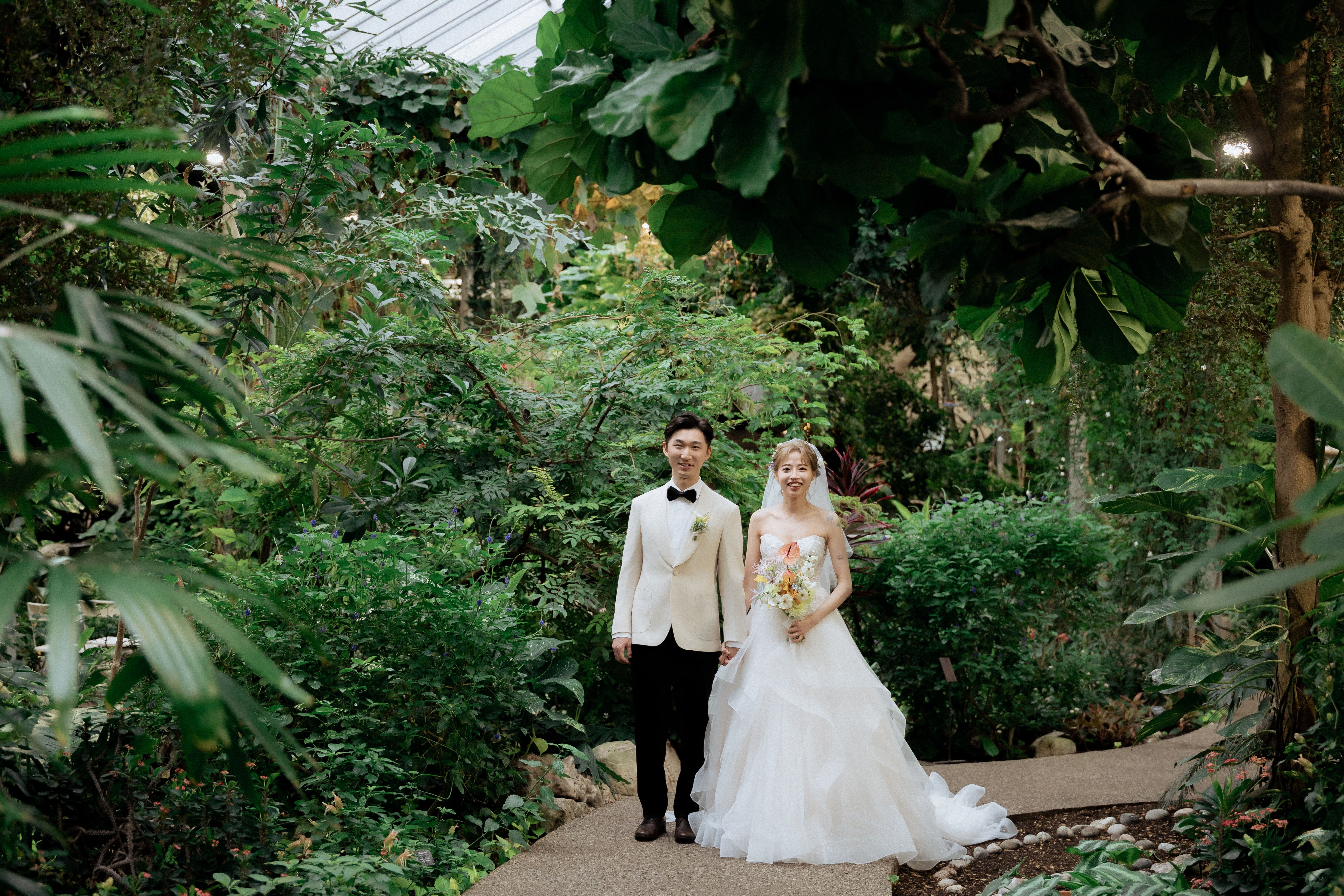Bride and groom on a winding path beneath layered foliage and a glass roof at Cambridge Butterfly Conservatory.