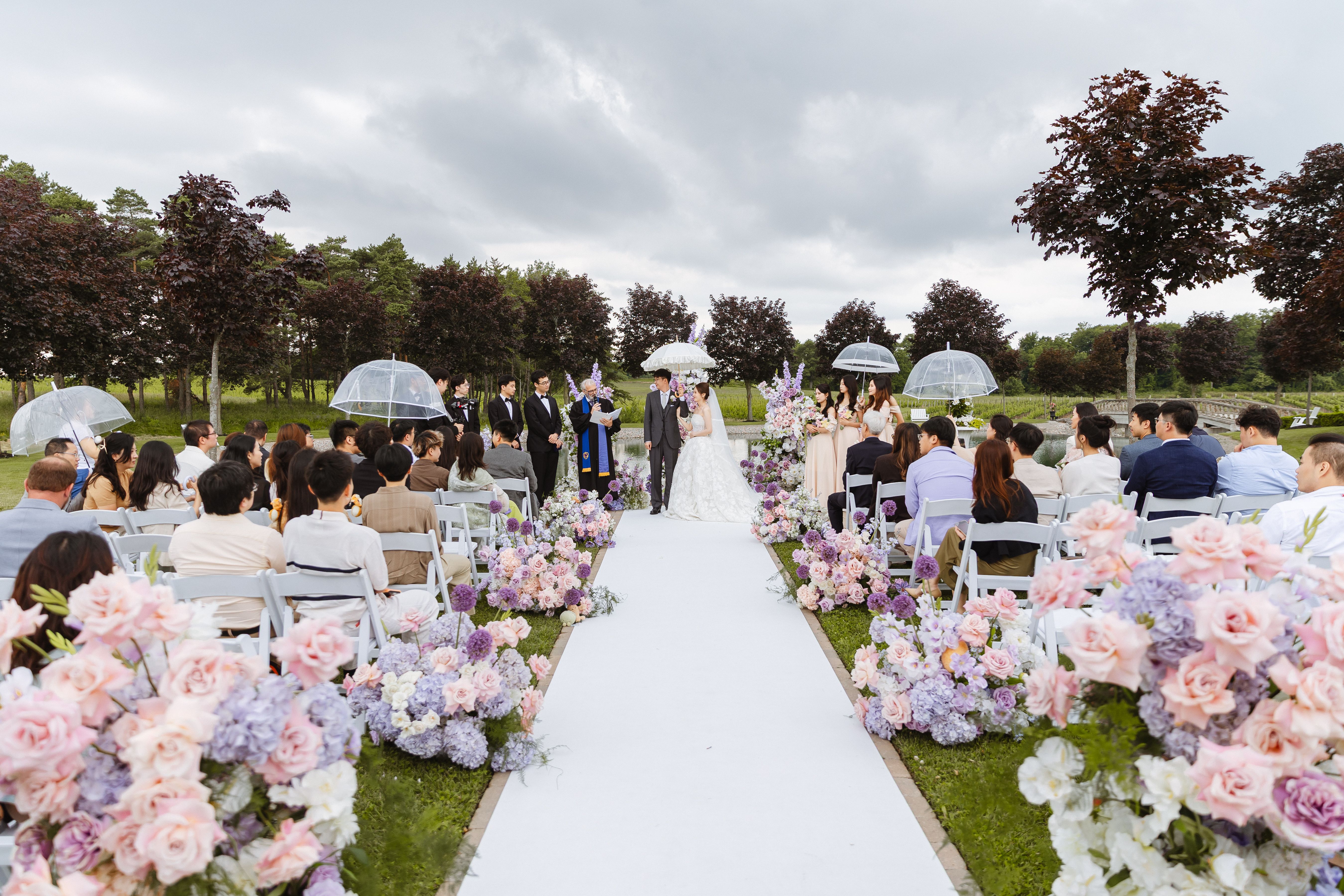 Guests seated beside a flower-lined outdoor aisle at Lincoln Estates, with the couple at the front and clear umbrellas beneath a cloudy sky.