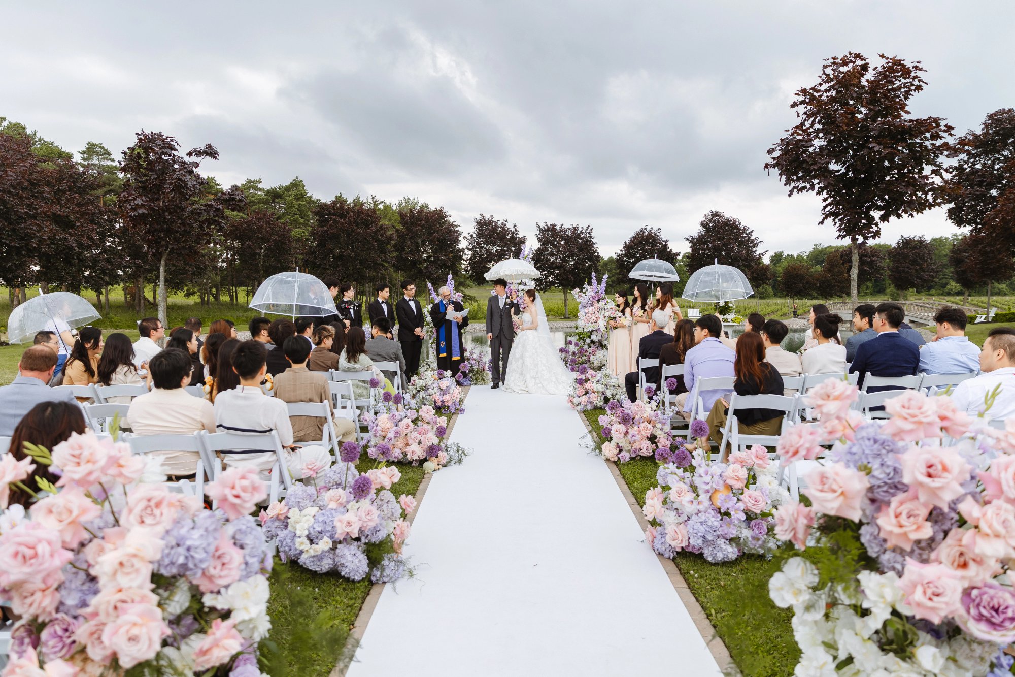 Guests seated beside a flower-lined outdoor aisle at Lincoln Estates, with the couple at the front and clear umbrellas beneath a cloudy sky.