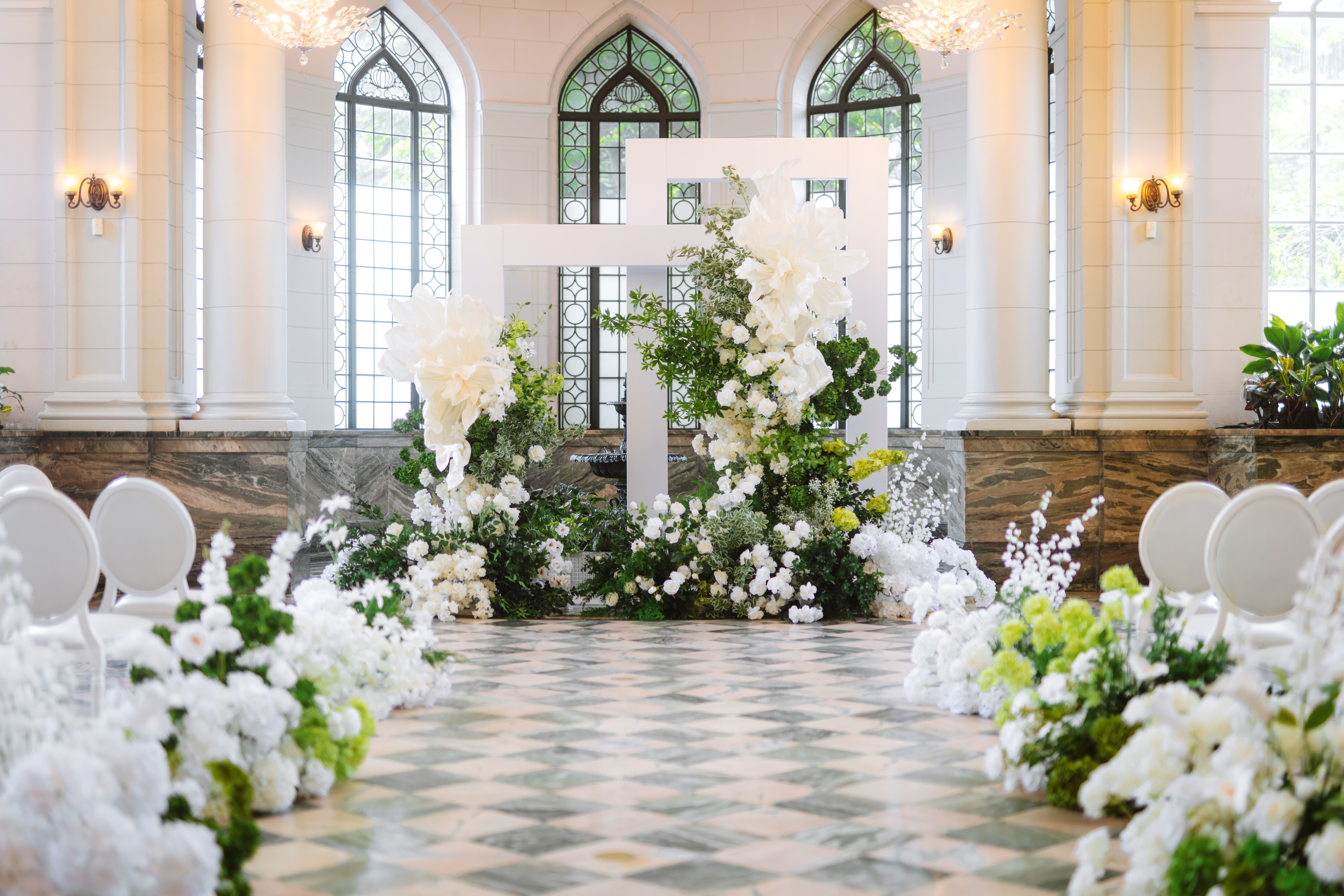 White flowers and greenery line a tiled ceremony aisle beneath pointed arched windows at Casa Loma.