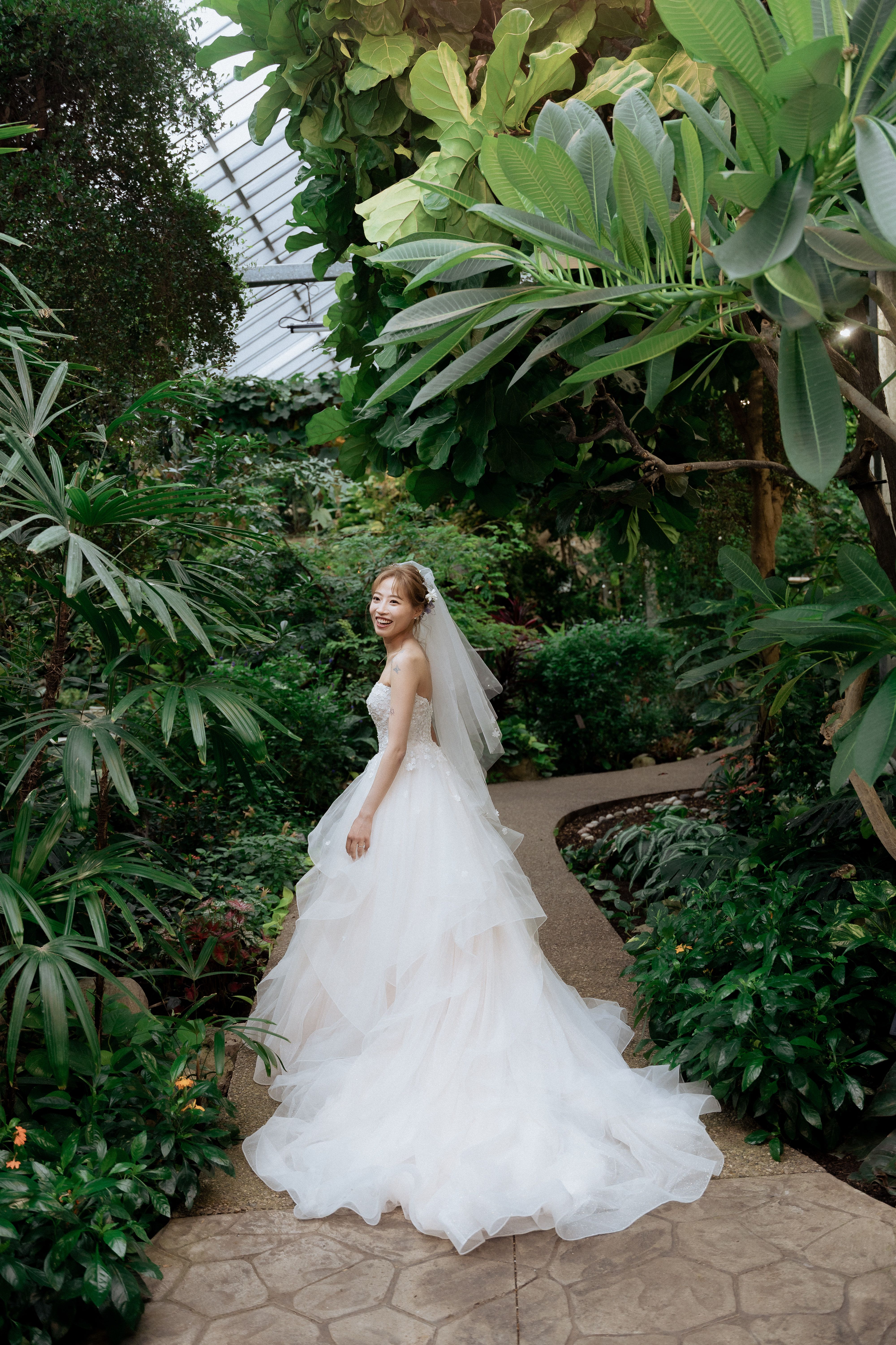 Bride looking back in a layered white gown on a plant-lined path at Cambridge Butterfly Conservatory.