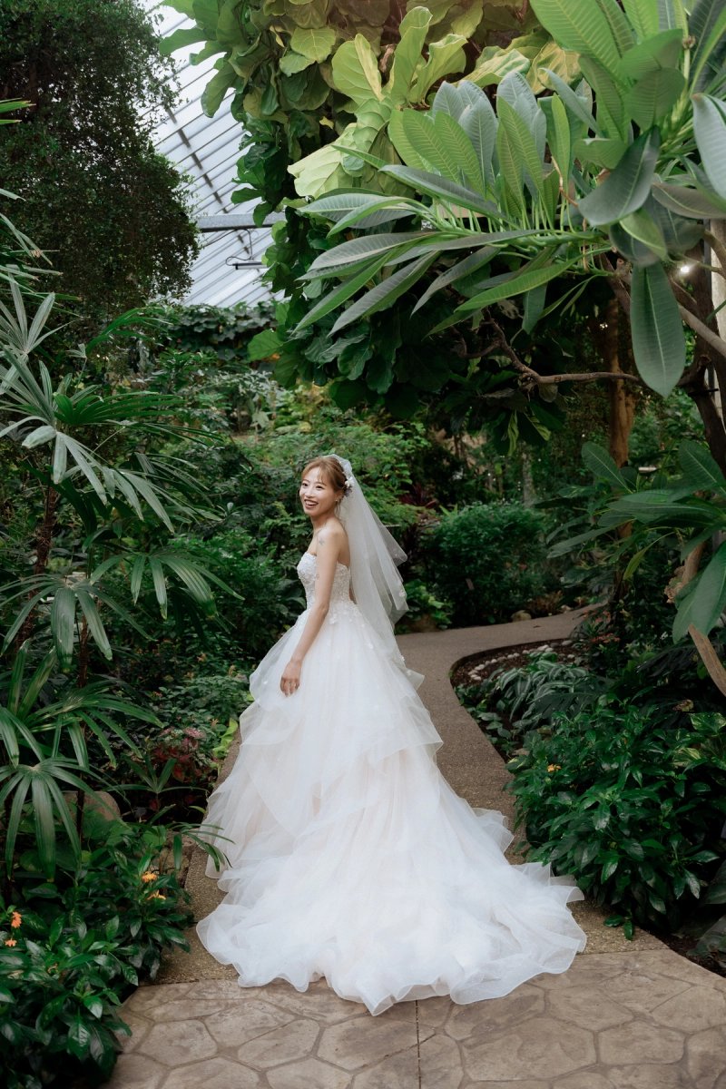 Bride looking back in a layered white gown on a plant-lined path at Cambridge Butterfly Conservatory.