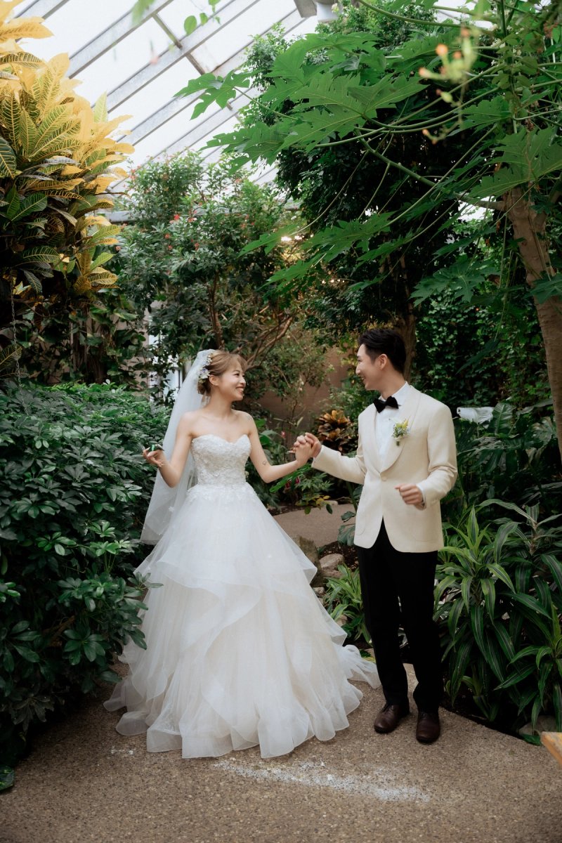 Bride and groom smiling at each other with hands joined on a leafy path at Cambridge Butterfly Conservatory.