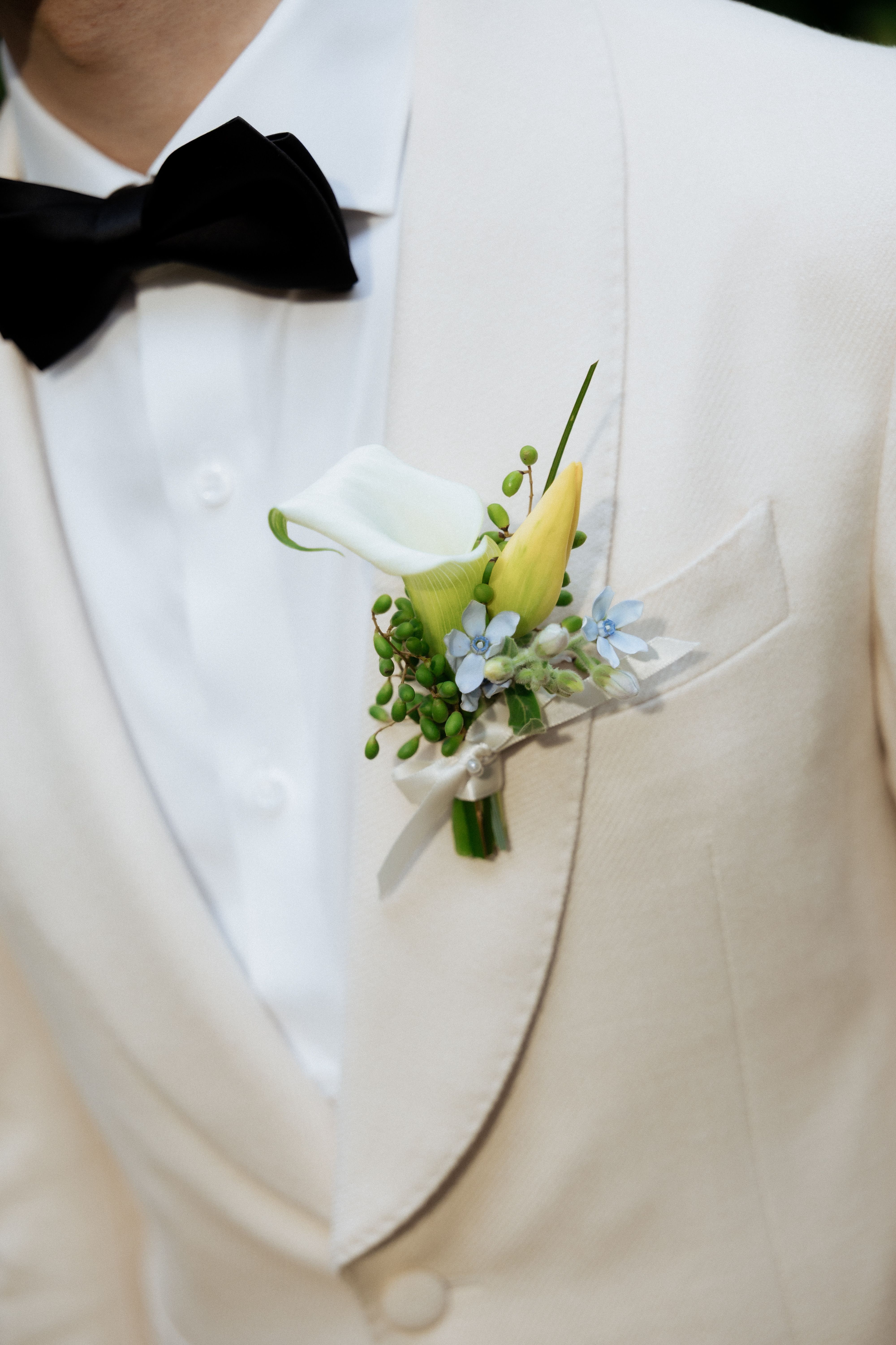 Pale yellow and blue boutonniere on the groom's ivory jacket beneath a black bow tie at Cambridge Butterfly Conservatory.