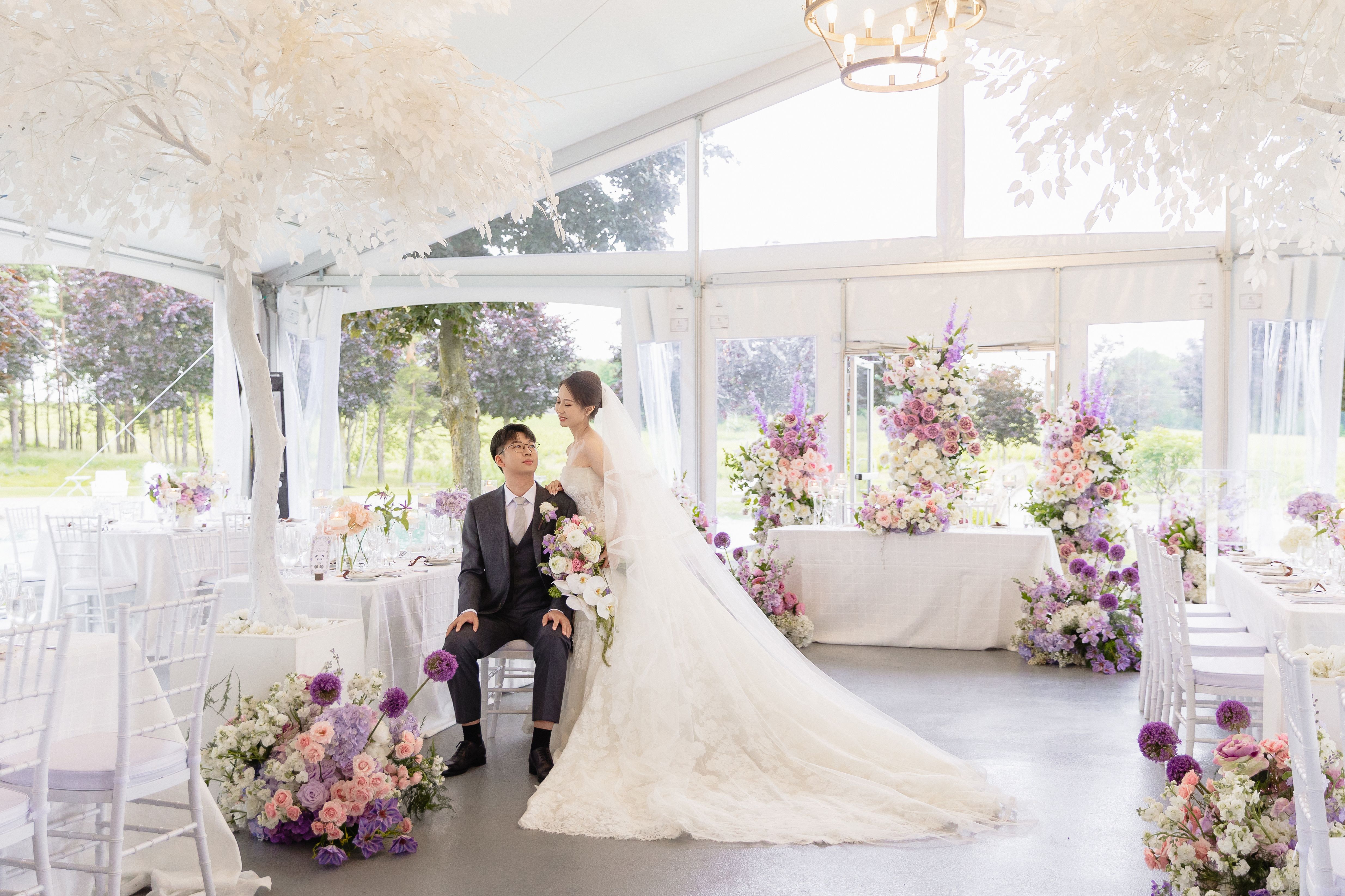A seated groom beside a standing bride in the Lincoln Estates reception tent, surrounded by flowers and white decorative trees.
