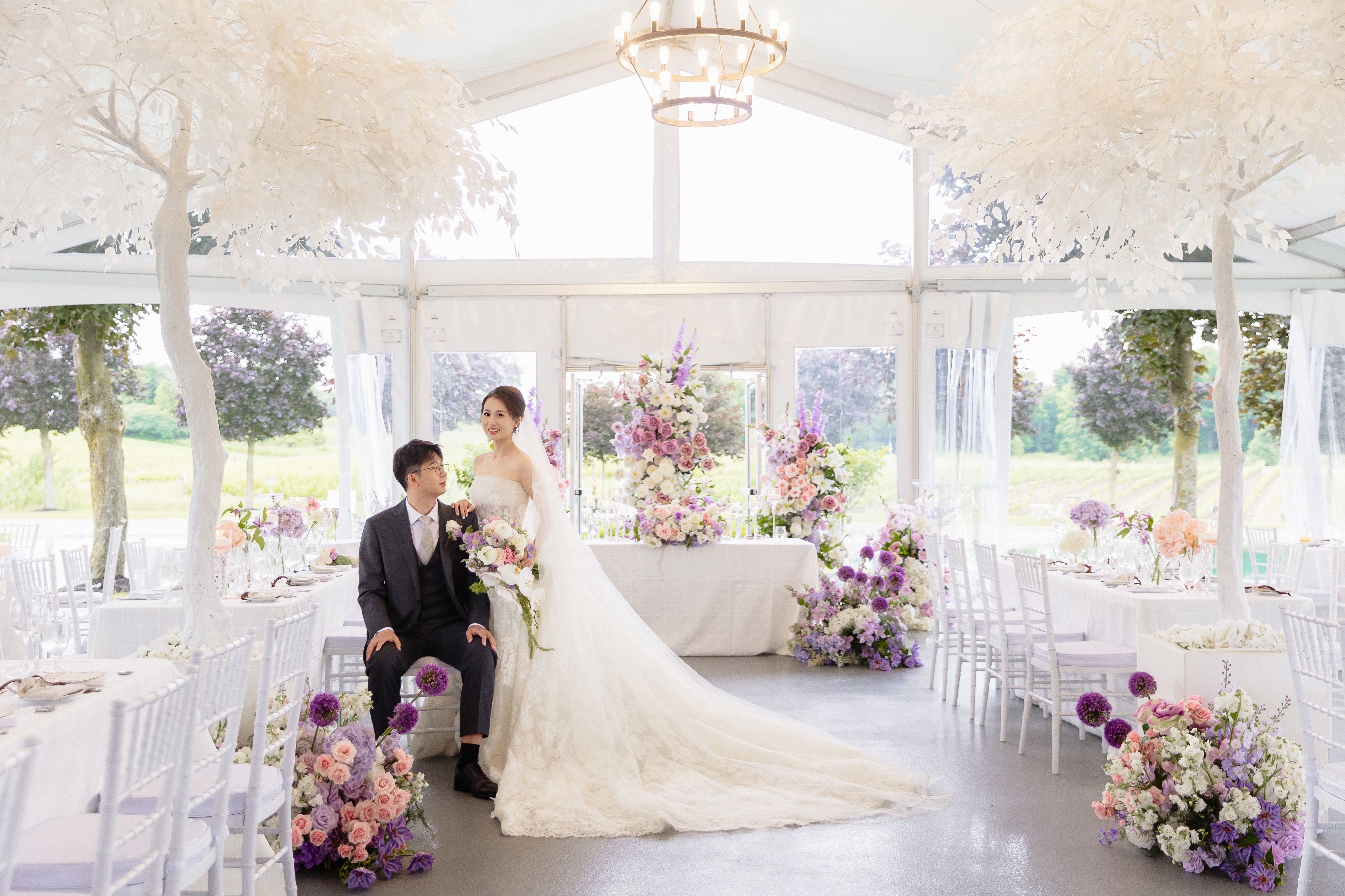A bride standing beside a seated groom in the Lincoln Estates reception tent, between white decorative trees and rows of white chairs beneath a chandelier.