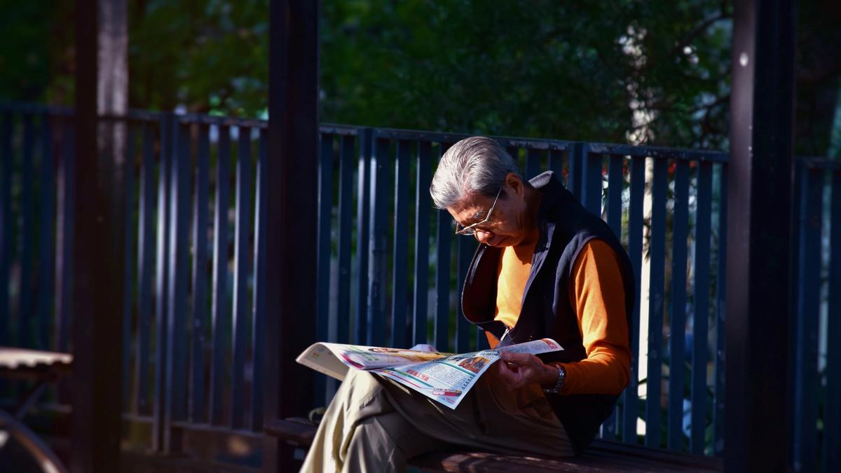 Eldre mann med orange genser og svart vest sitter på en benk i sola å leser avisen. Illustrasjonsbilde tatt av Mark Hang Fung So fra Unsplash