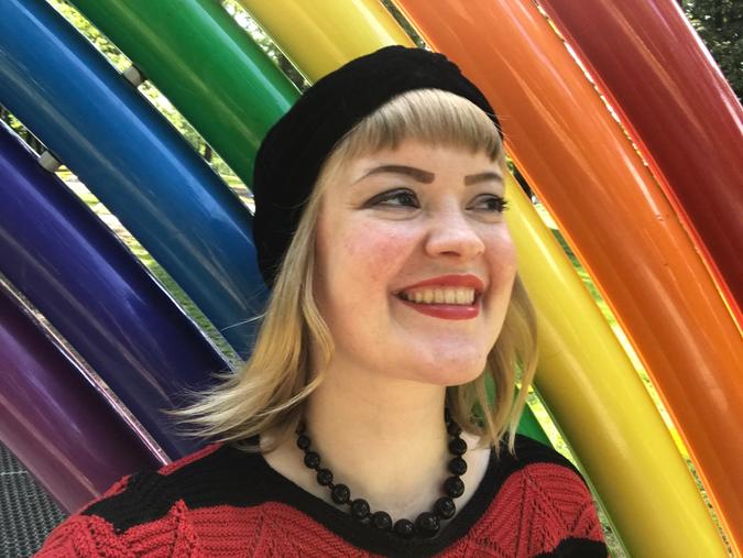 Jente med blondt hår og svart lue. Hun står foran en regnbue statue.