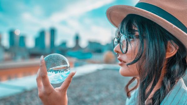 Illustrasjonsbilde av en kvinne med briller og hatt som holder en gjennomsiktig ball som reflekterer bybilde i bakgrunnen. Bilde tatt av Maryna Yazbeck fra Unsplash