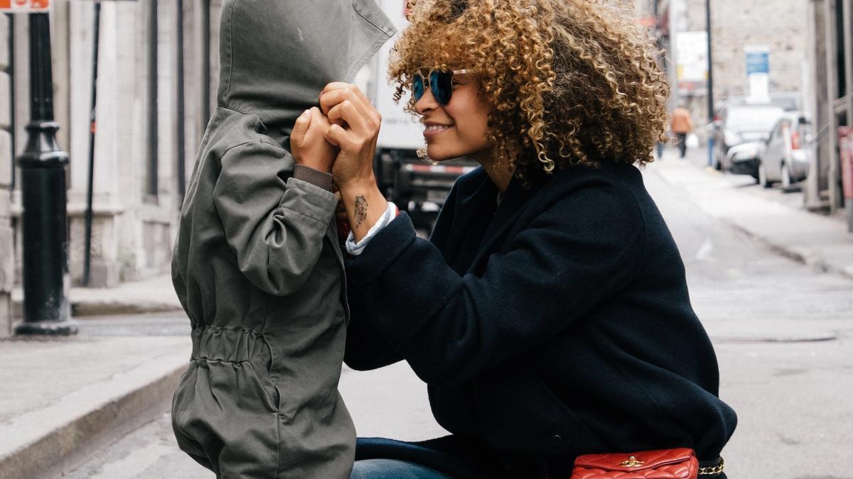 Illustrasjonsbilde av en mor med svart kåpe og solbriller som holder hendene til et lite barn i grå dress og smiler. Bakgrunnen er et bybilde. Illustrasjonsbilde tatt av Sai De Silva fra Unsplash