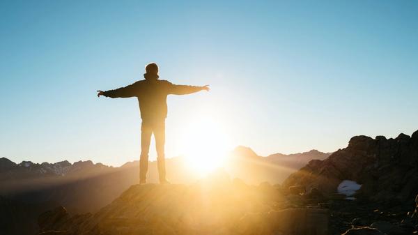 Illustrasjonsbilde av en mann som har armene ut mens han står å ser utover et fjell-landskap og en solnedgang. Bildet er tatt av Pablo Heimplatz fra Unsplash