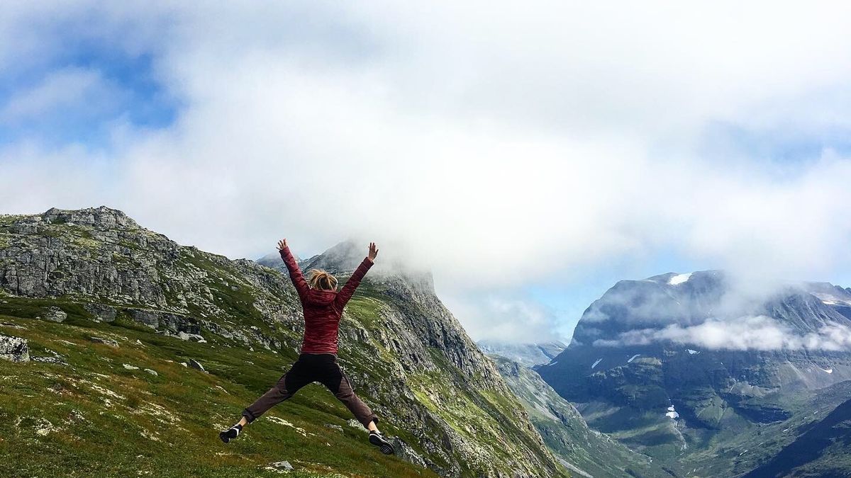 Kvinne som hopper på fjellet. Blå himmel med skyer bak grønne fjell. Bilde privat, Tine Norderhaug